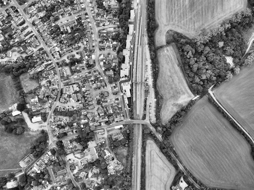 An aerial view of a rural landscape showing a large expanse of lush green fields divided by narrow dirt paths and low hedgerows. In the foreground, there is a farmstead with several farm buildings, in